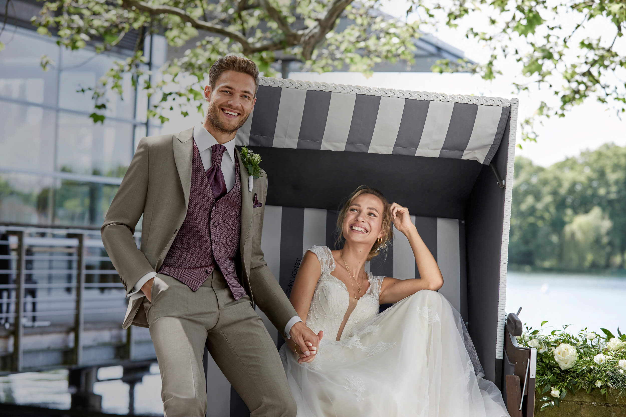 Ein Brautpaar welches in einem Strandkorb sitzt. Die Hochzeitsmode ist aus der aktuellen Kollektion von Wilvorst, die Braut- und Festmode von Lauretta Schmitz in der Naehe von Aachen führt.